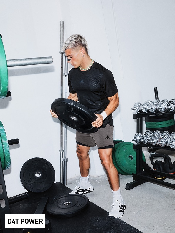 Una persona con una camisa y shorts negros está levantando pesas en un gimnasio, rodeada de varios equipos y máquinas para hacer ejercicio.