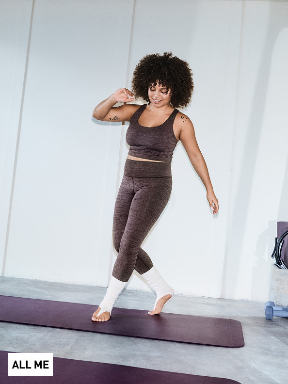 Una mujer de pelo rizado parada sobre un mat de yoga en un ambiente de estudio brillante y minimalista.