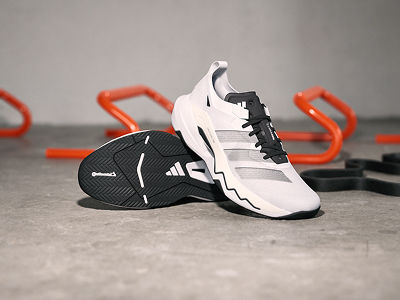 A pair of white and black Adidas athletic shoes resting on a grey concrete surface, with orange exercise equipment visible in the background.