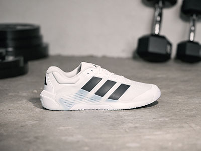 A pair of white and black Adidas trainers rests on a concrete floor, with a blurred background of exercise equipment visible.