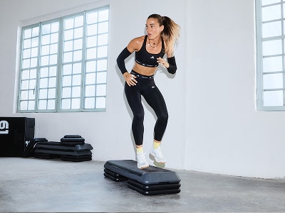 Una mujer vestida con ropa deportiva está de pie sobre una plataforma de vibración en una sala abierta y brillante con grandes ventanas.