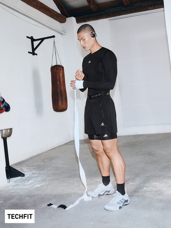 A person in black athletic clothing stands in a home gym setting, with a punching bag and other fitness equipment visible in the background.