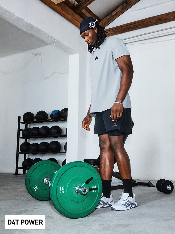 A person wearing a white shirt, black shorts, and trainers is standing in a gym setting, with weight plates and a barbell visible in the foreground.