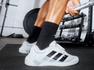 The image shows a person's lower body wearing white trainers and black socks, standing in a gym setting with weight equipment visible in the background.