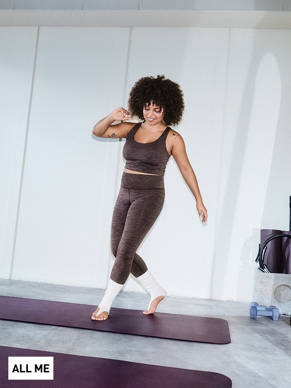 A woman with curly hair stands on a yoga mat in a minimalist studio setting, wearing athletic clothing and appearing to be in a yoga or exercise pose.