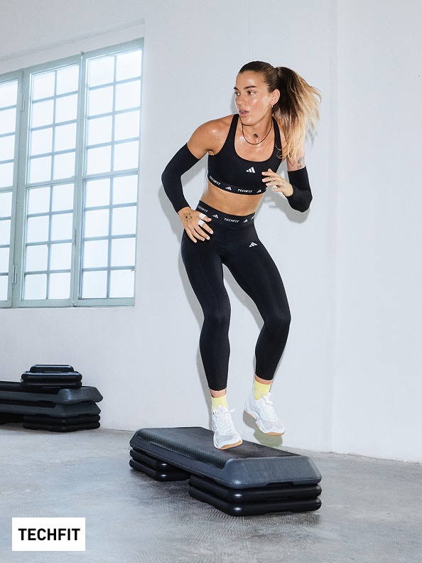 A woman in athletic wear is standing on a black exercise platform in a bright, windowed room.