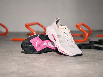 The image shows a pair of white and pink athletic shoes with orange accents, resting on a grey surface alongside a black and pink exercise mat.