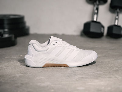 A pair of white trainers sits on a concrete floor, with a blurred background of exercise equipment visible.