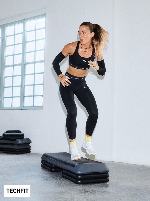 Una donna in abbigliamento sportivo è in piedi su una piattaforma di fitness nera in una stanza luminosa e rifinita da finestre.