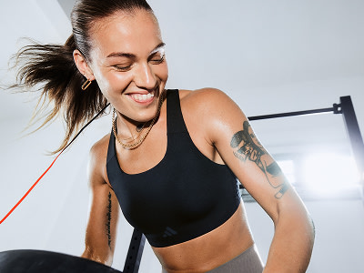 Una mujer sonriente con un top deportivo negro de pie en un gimnasio, con equipo para ejercitarte visible en el fondo.
