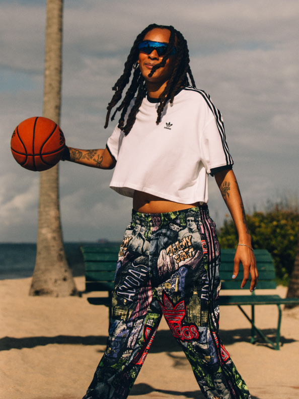 A person with dreadlocks wearing sunglasses and a white shirt stands on a beach, holding a basketball, with palm trees and a body of water visible in the background.