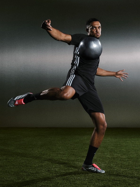 A man in a black athletic outfit is jumping and reaching out with one arm on a dimly lit sports field.