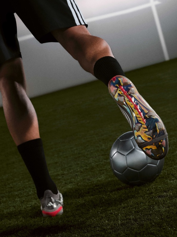 A person's lower body and feet are visible, wearing colorful athletic shoes and socks, standing on a soccer ball on a grassy field.