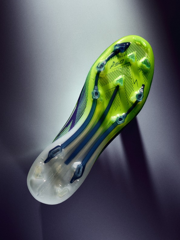Ein leuchtend grün-blauer Fußballschuh mit cleanem, aerodynamischem Design hebt sich von einem dunklen, schattigen Hintergrund ab.