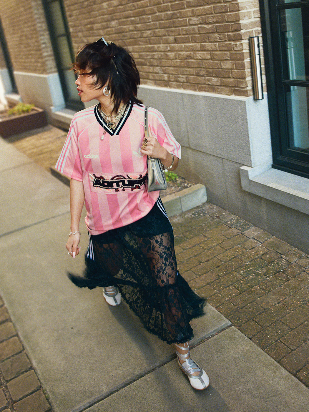 A person wearing a pink and black striped shirt, a black lace skirt, and white sneakers stands in front of a brick building.