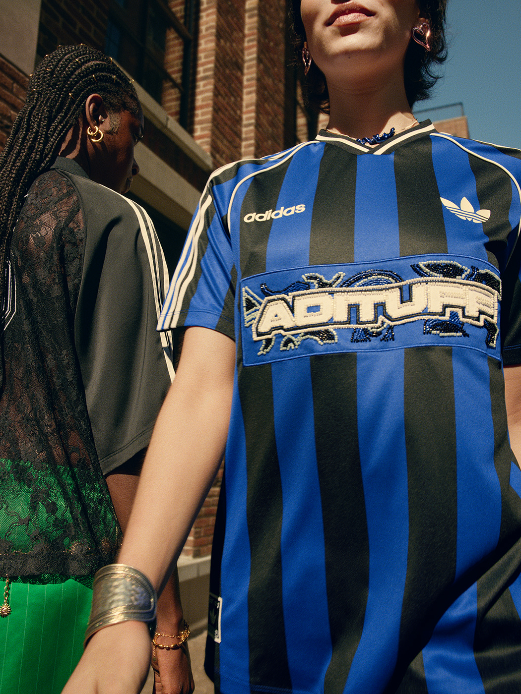The image depicts two individuals wearing sports jerseys, one in blue and white stripes and the other in a dark color, standing in front of a brick building.