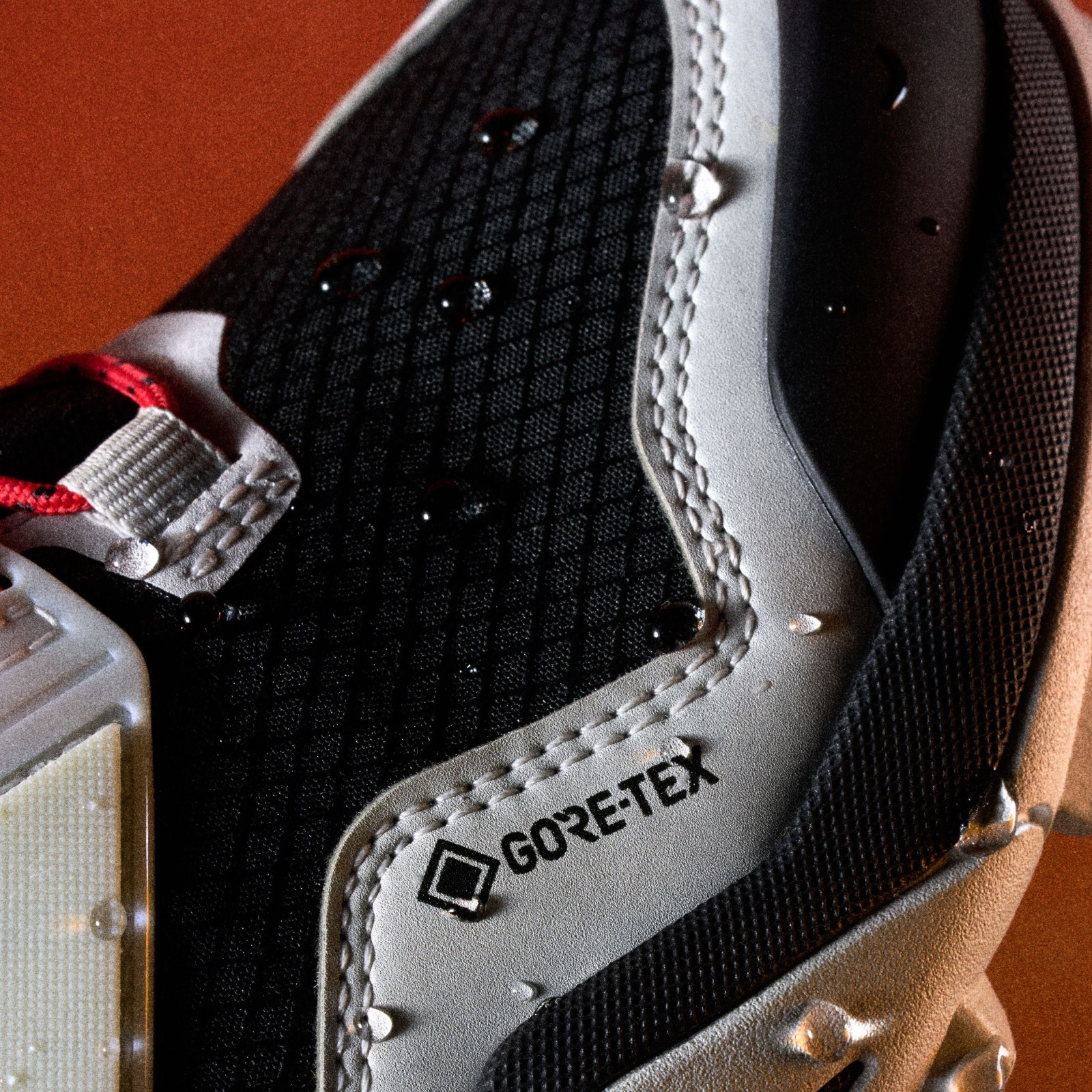 A close-up view of a black and white athletic shoe with the GORE-TEX logo prominently displayed, set against a reddish-orange background.
