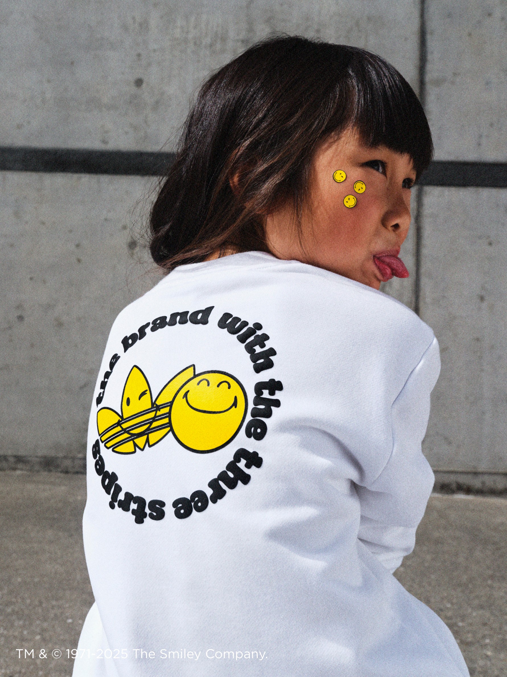 A young person wearing a white sweatshirt with a graphic design of smiling faces, standing in front of a concrete wall.