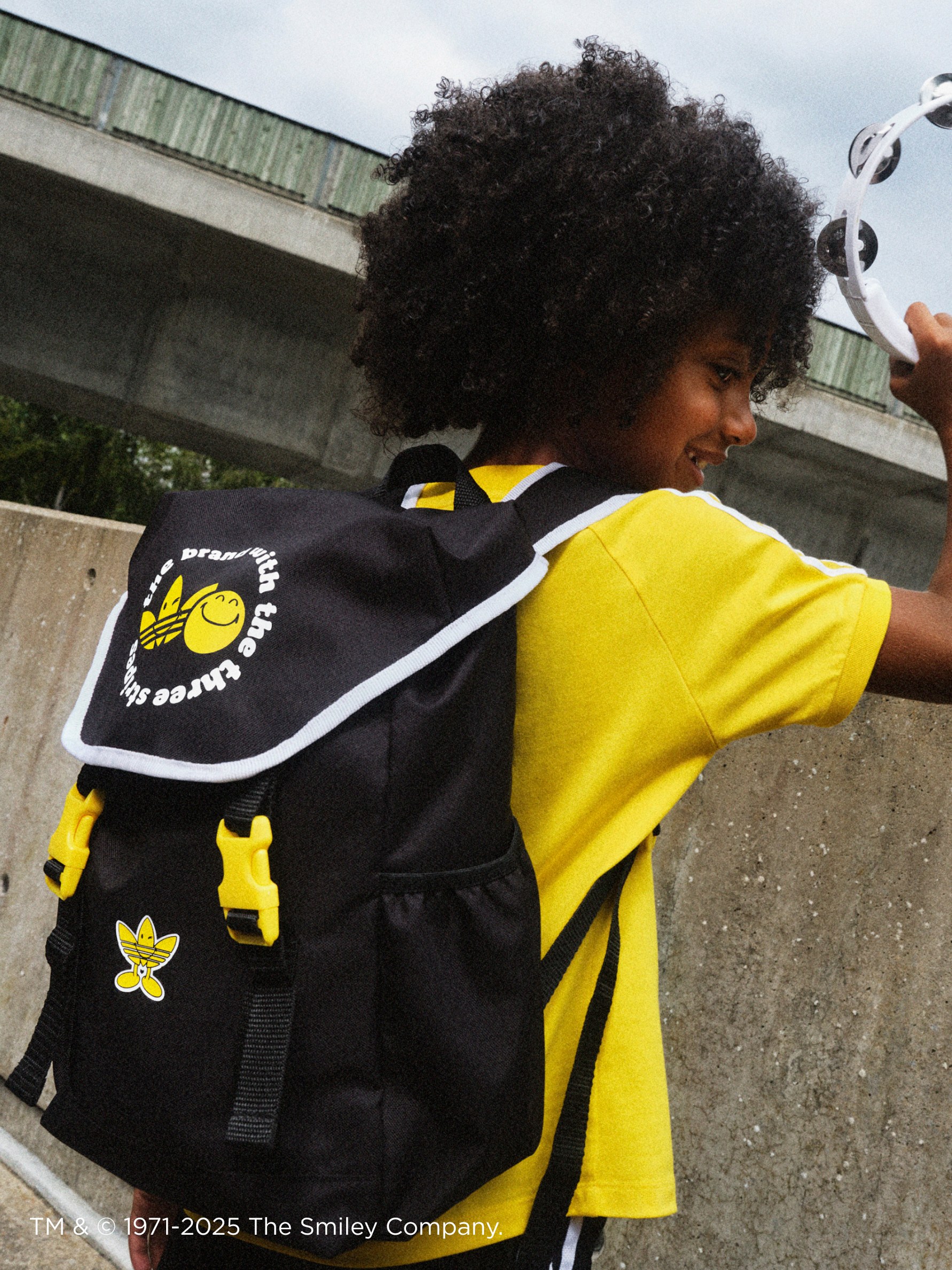 A person with curly dark hair wearing a black backpack with yellow designs stands in front of a concrete structure.