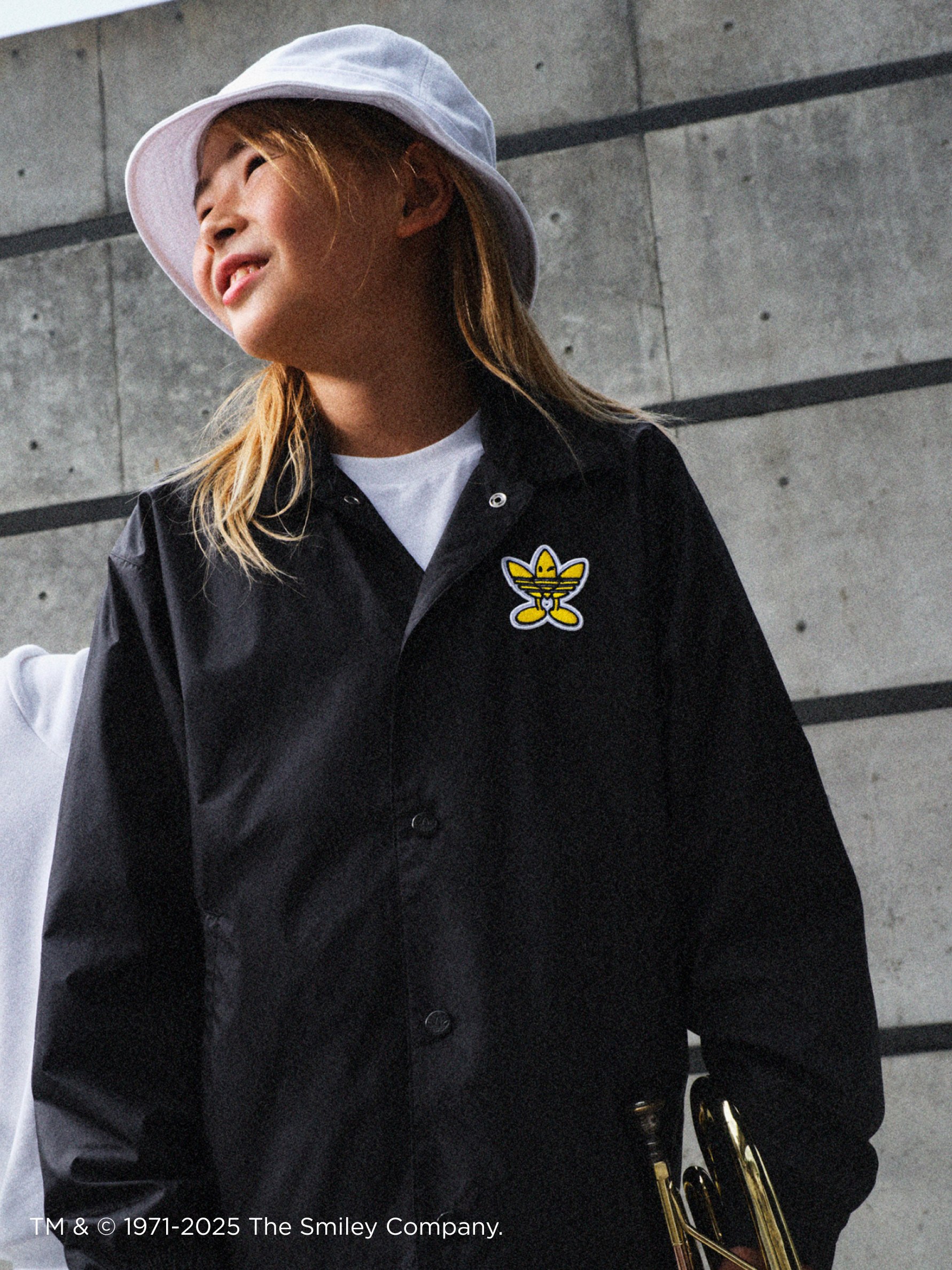 A young person wearing a black jacket with a yellow emblem, standing in front of a concrete wall.