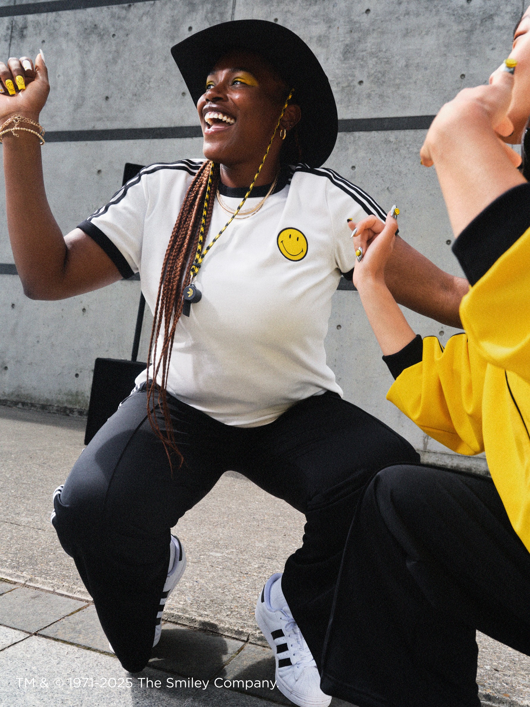 A smiling person wearing a black hat, white t-shirt with a smiley face, and black pants is seated on the ground in front of a concrete wall.