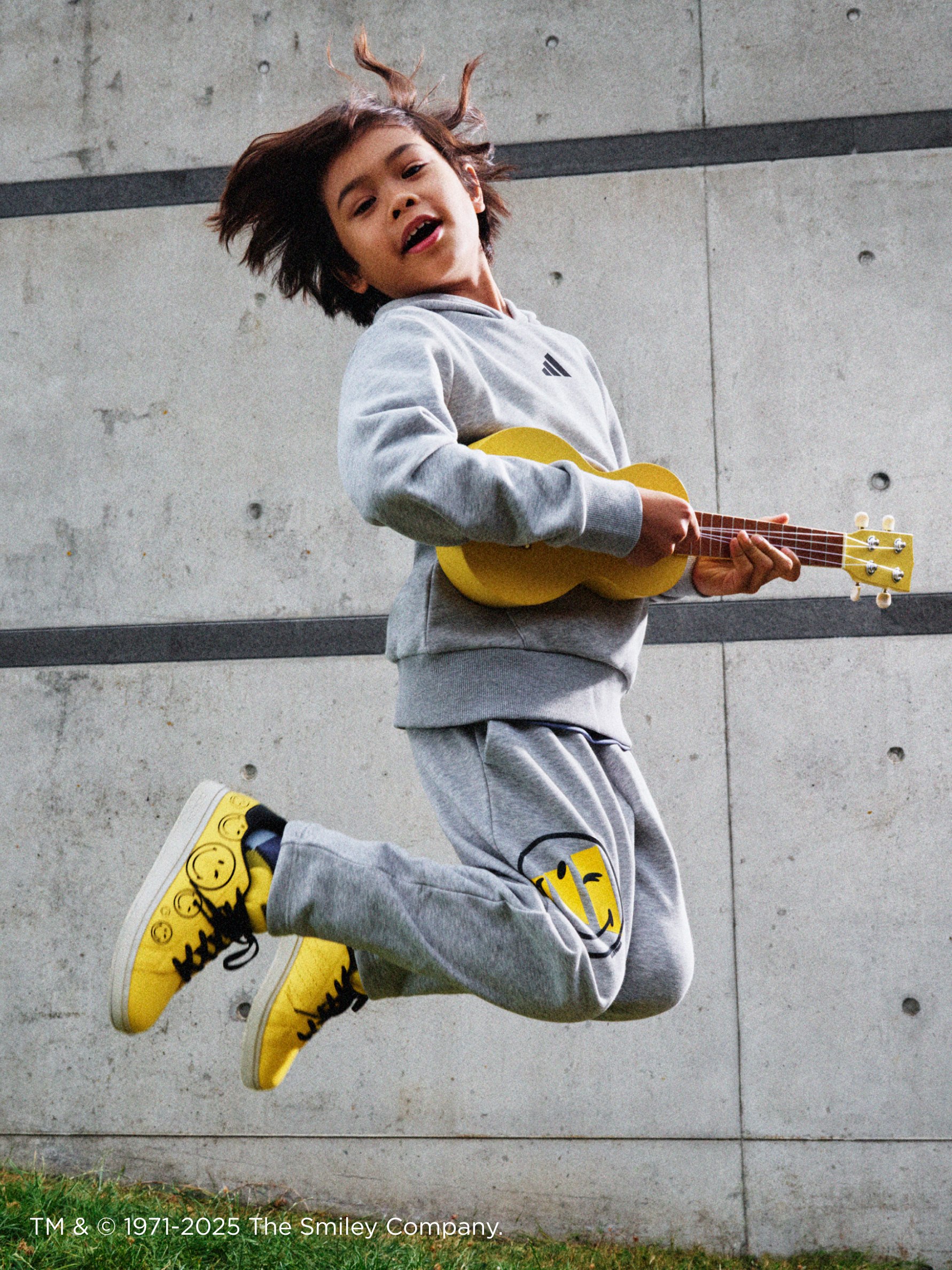 A person in a gray sweatsuit is jumping and playing a guitar against a concrete wall backdrop.