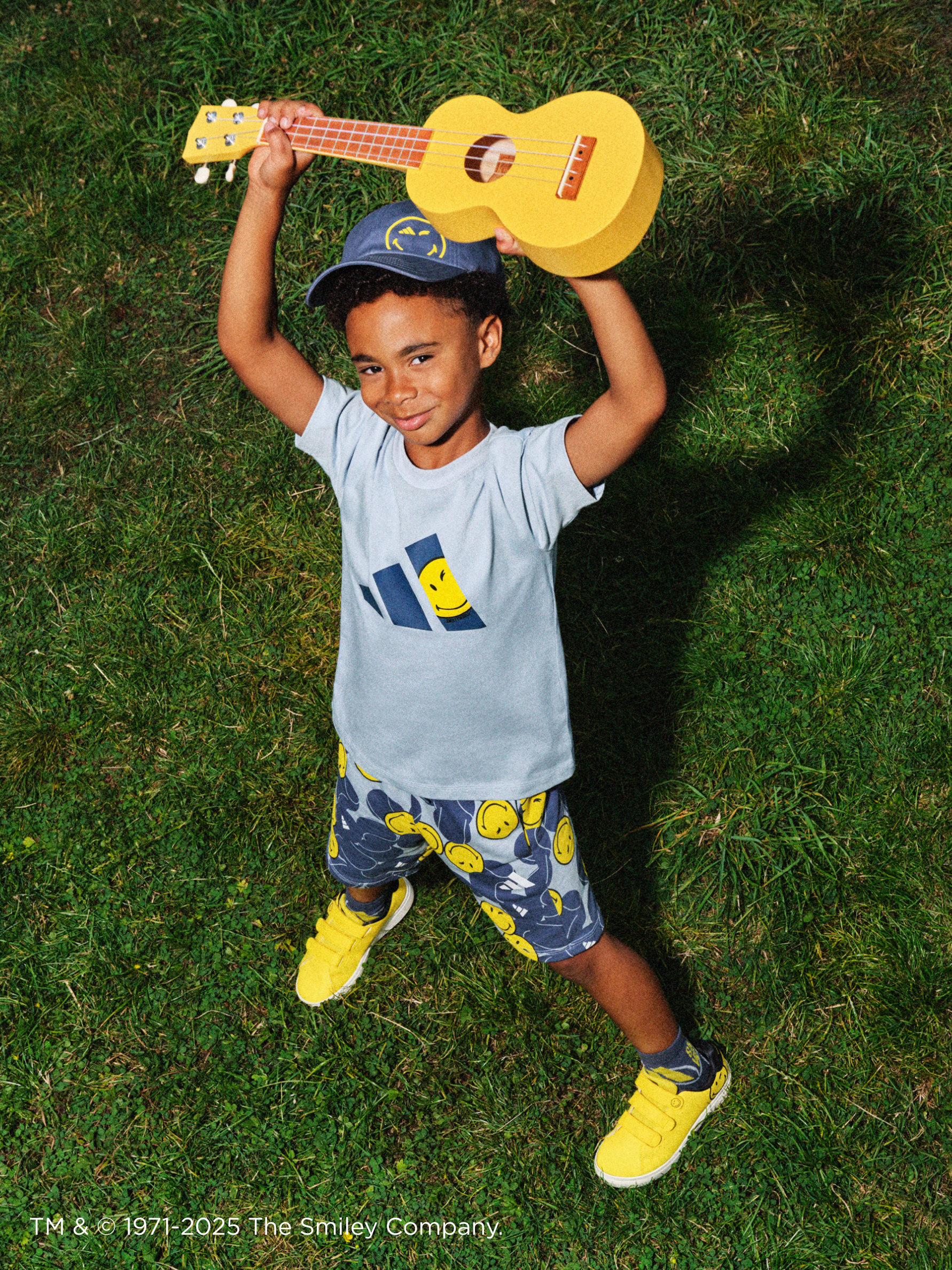 A young child wearing a gray t-shirt and blue shorts is holding a large yellow ukulele-like toy instrument while lying on a grassy field.