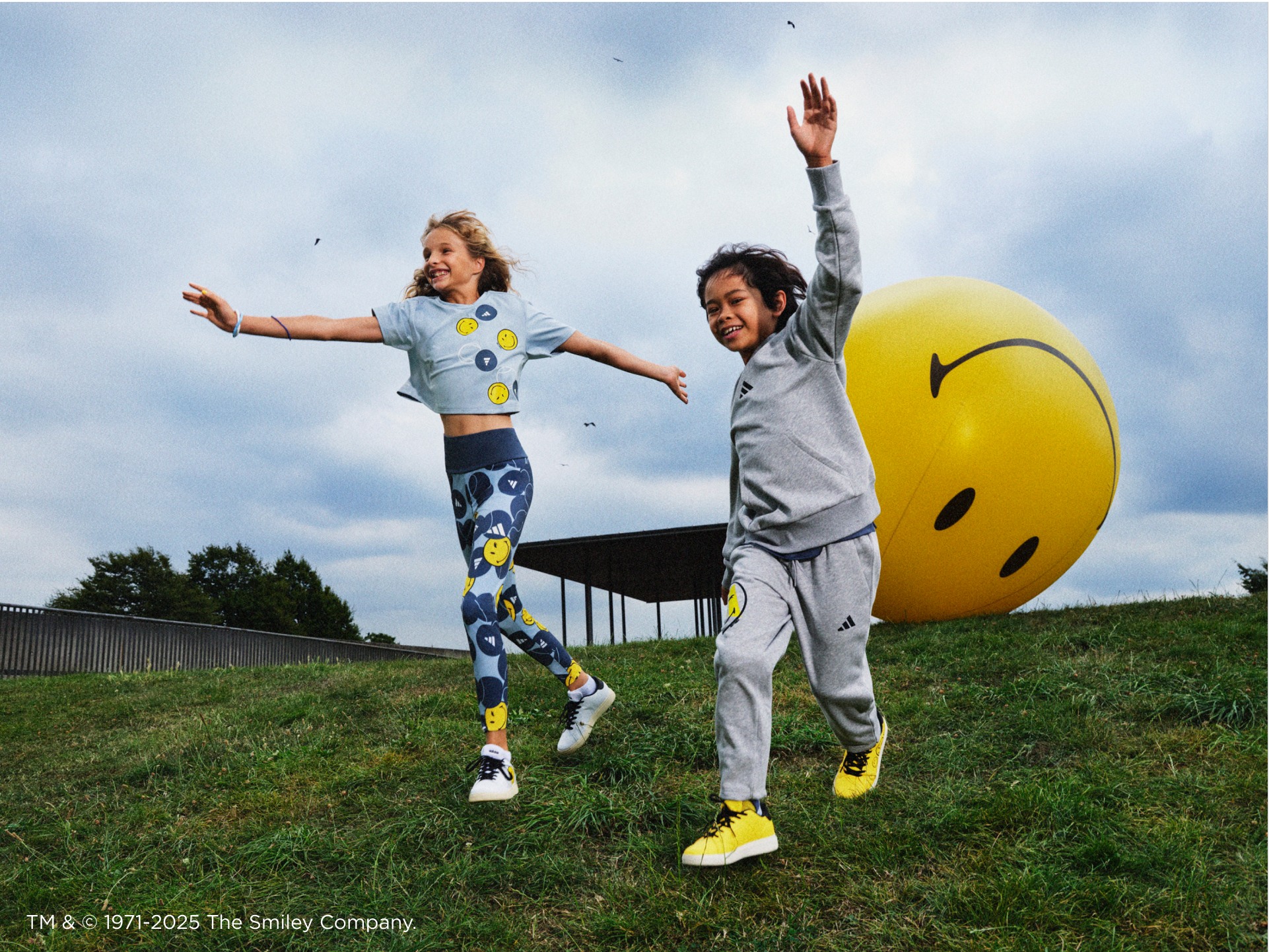 Two kids wearing adidas | Smiley sportswear run down a hill, followed by a giant yellow inflatable ball.
