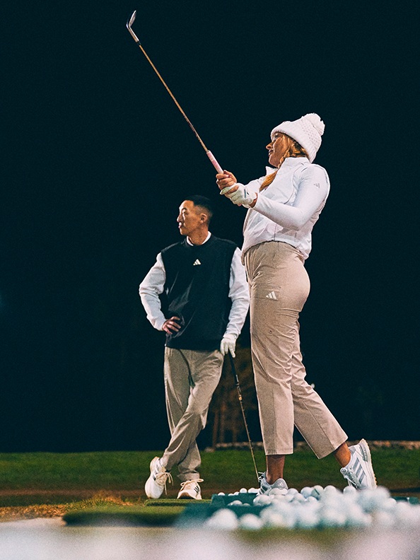 Duas pessoas, um homem e uma mulher, estão de pé num campo de golfe à noite, com a mulher a segurar um clube de golfe e o homem ao seu lado.