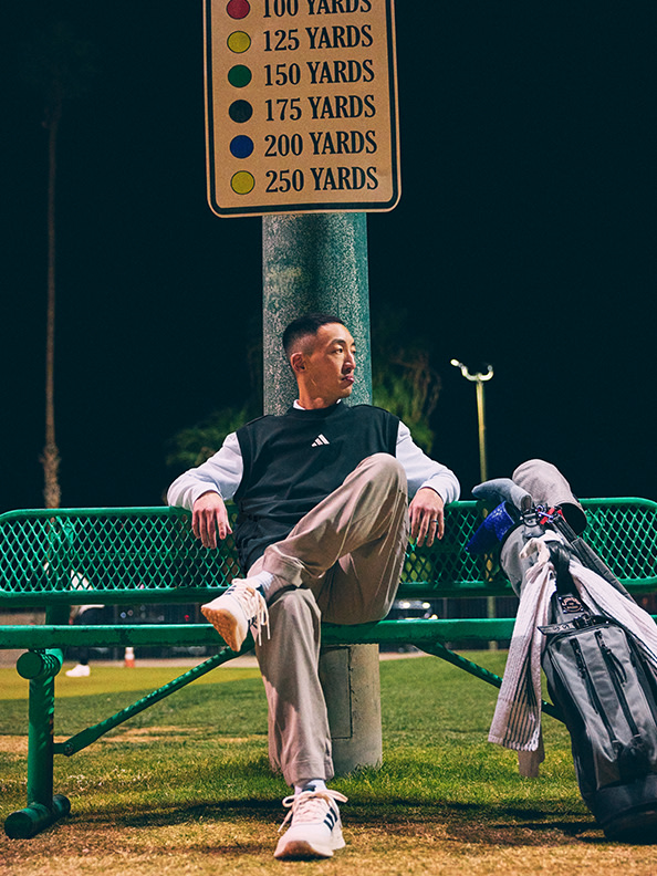 A man in casual attire sits on a bench near a yardage marker on a sports field, with golf bags visible in the background.