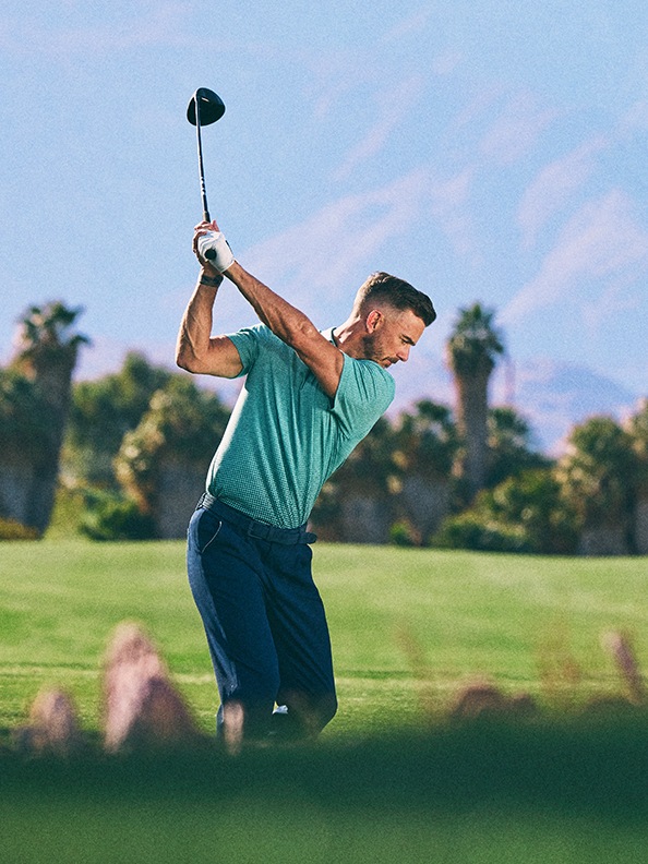 Um homem de camisa verde está a balançar um clube de golfe num campo de golfe exuberante e relvado com palmeiras ao fundo.