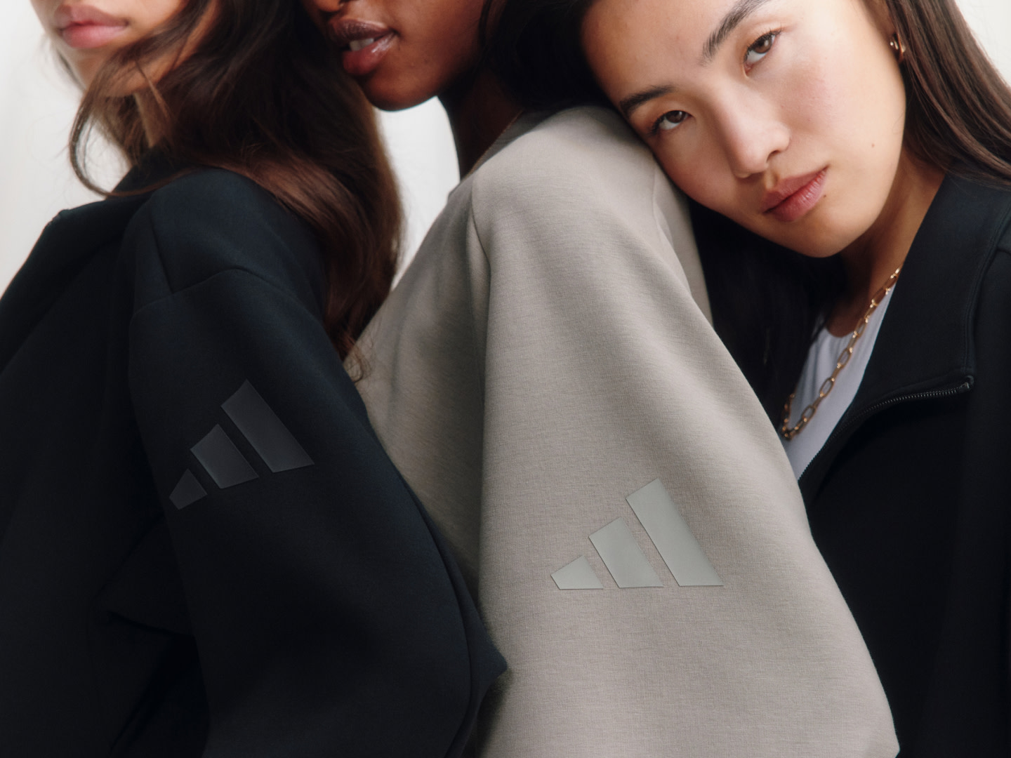 Three young women with dark hair wearing black and gray clothing, standing close together against a plain background.