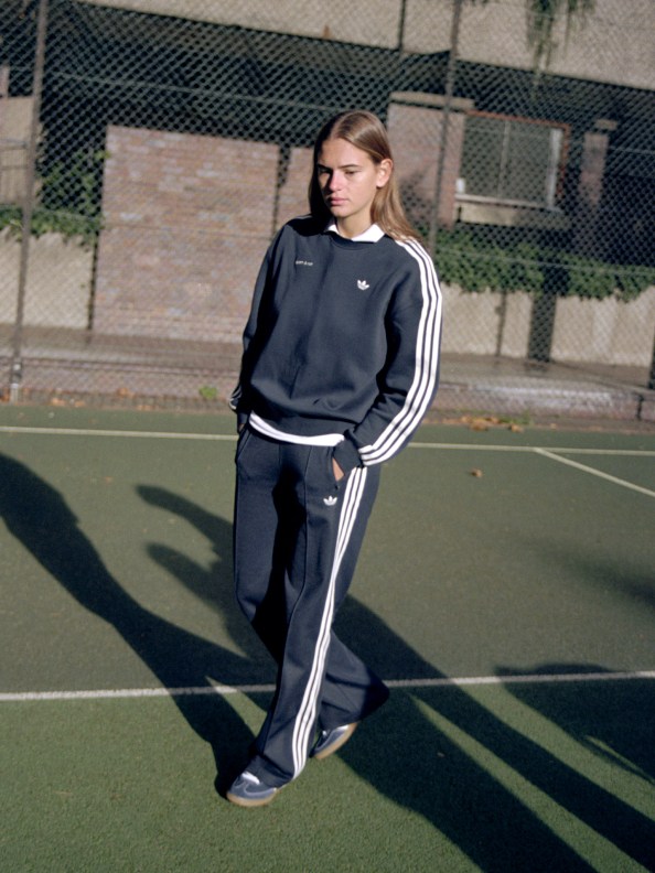 A blonde woman wears a dark blue adidas x Sporty & Rich crewneck, track pants, and Handball Spezial shoes while strolling a tennis court.