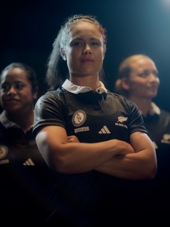 A young woman in a black sports jersey stands with her arms crossed, surrounded by other people in the background.