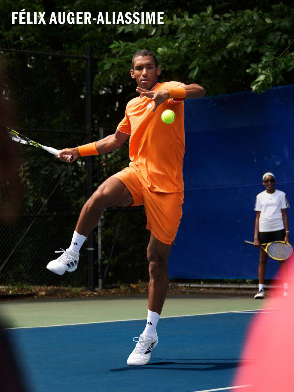 A man in an orange shirt and white shorts is playing tennis on a tennis court, with another person visible in the background.