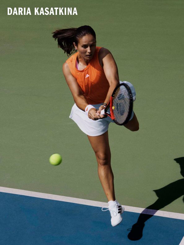 A female tennis player in an orange top and white shorts is shown in mid-swing, preparing to hit a tennis ball on a tennis court with a green surface.