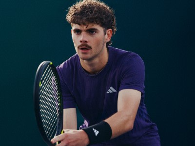 En ung man med lockigt hår iklädd en lila atletisk tröja och med ett tennisracket står på en tennisplan mot en mörkgrön bakgrund.