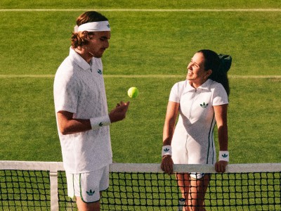 Twee personen, een man en een vrouw, staan op een tennisbaan en gaan in gesprek. De achtergrond is een grasveld, wat een outdoor-tennisfaciliteit suggereert.