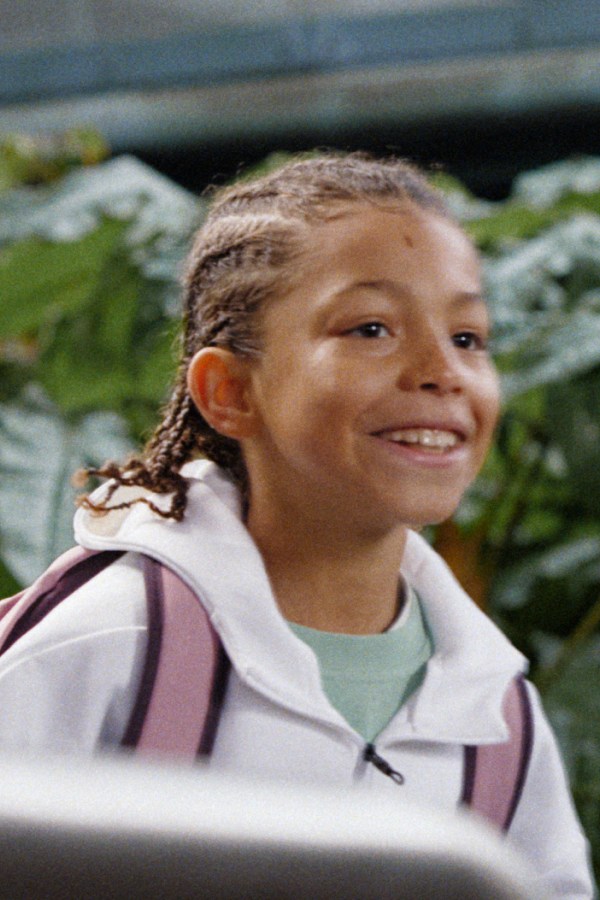 A young girl with braided hair is smiling brightly, standing in front of a lush, green background.