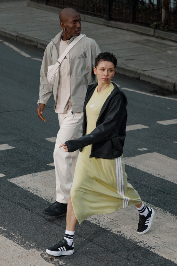 Two individuals, a man and a woman, are walking together on a street. The man is wearing a light-colored outfit, while the woman is wearing a yellow dress and sneakers.