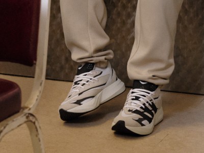 The image shows a pair of white and black sneakers on a person's feet, with the person wearing beige pants in a room with a patterned carpet.