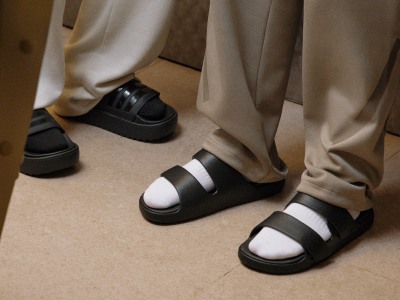 The image shows a pair of black and white sandals on a wooden floor, with beige curtains visible in the background.