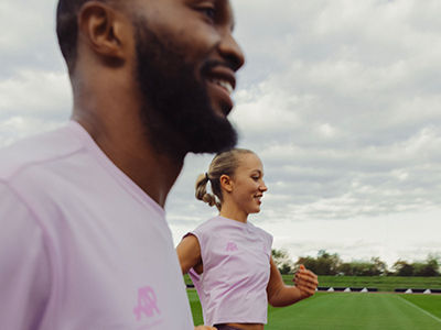 A tall, bearded man in a pink shirt stands in the foreground, with a young girl in a pink shirt visible behind him on a grassy field.