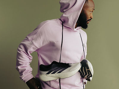 A person wearing a pink hooded sweatshirt stands against a neutral green background, with their hands resting on their waist.