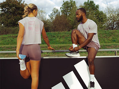 The image shows two people, a woman and a man, standing on a platform in a grassy outdoor setting with trees in the background.