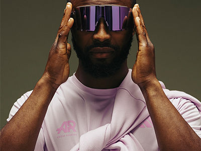 A man with a beard wearing a pink shirt and purple sunglasses, with his hands on his face against a plain background.