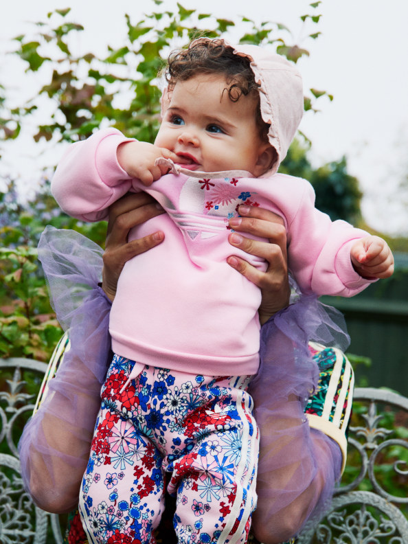 A young child, dressed in a pink jumper and floral pants, sits in a garden surrounded by lush greenery and flowers.