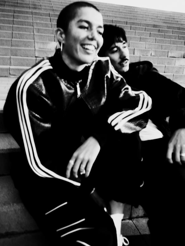 Two individuals, a man and a woman, are seated together in a casual, relaxed pose against a brick wall backdrop.