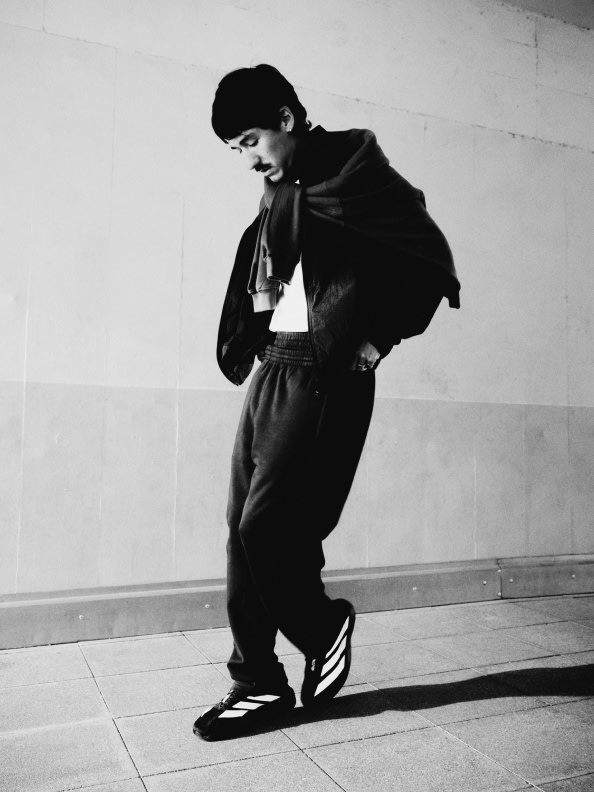 A person wearing a dark outfit and shoes stands in a plain, white-walled room.