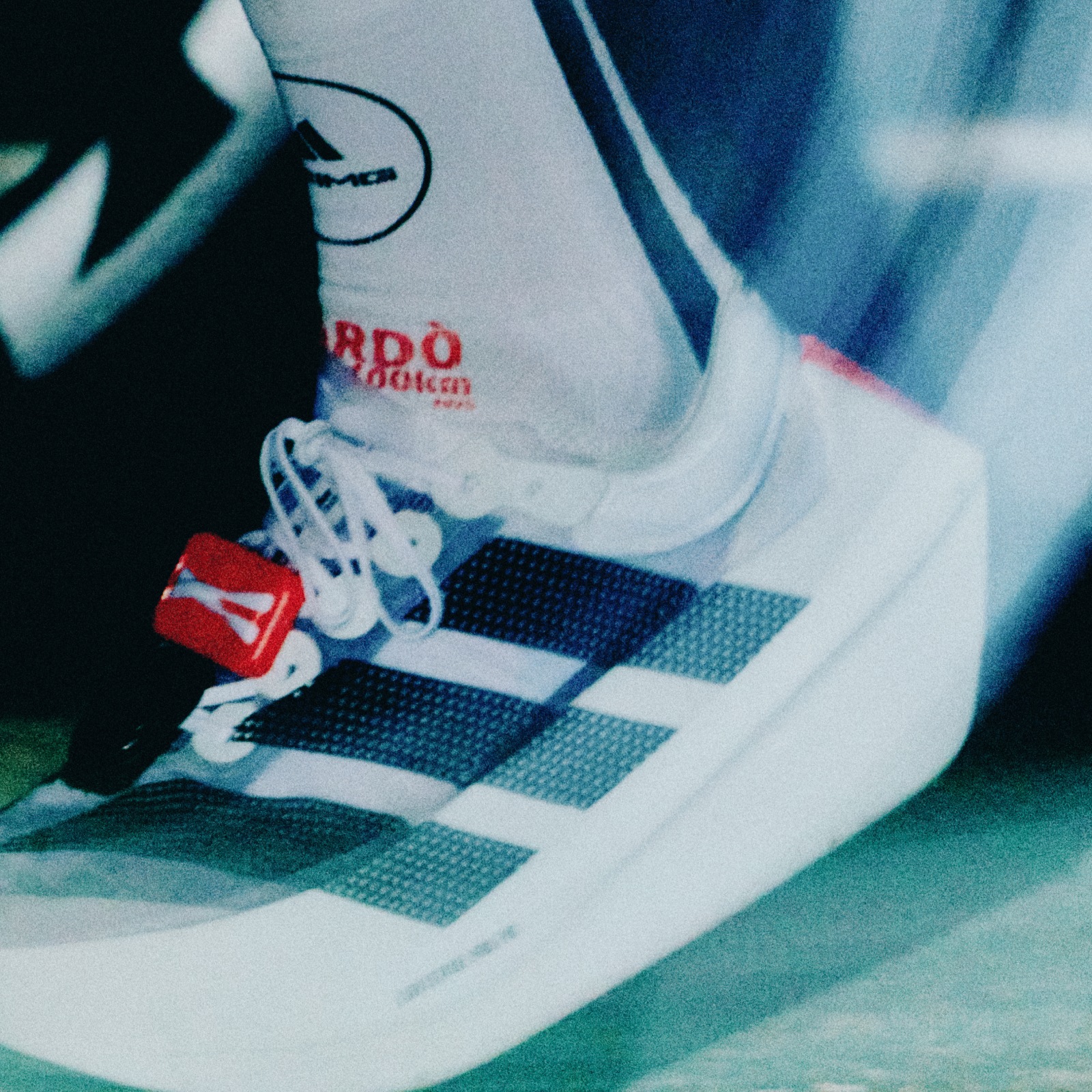 The image shows a close-up view of a person's foot wearing a white sneaker with the Adidas logo visible. The background appears to be a blurred, dark surface.